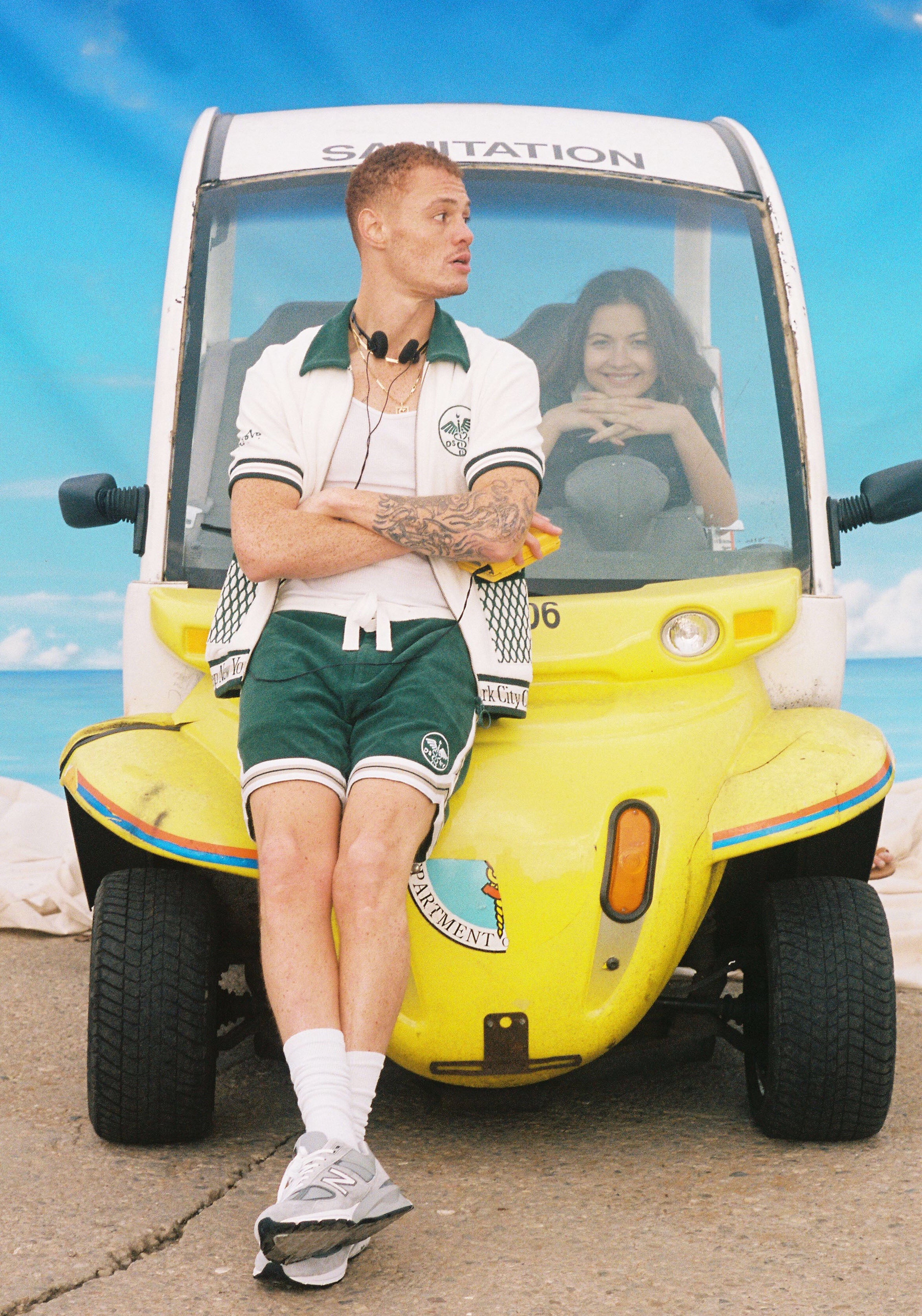 Man wears full terry cloth set and leans agaiinst Department of sanitation car while woman smiles in driver's seat