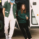 Female model sitting in door of garbage truck in DSNY sweater next to male model looking down in dsny hawaiian and hat