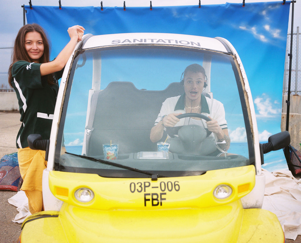 Male model mock driving the sanitation car in the DSNY cabana while a female model in the DSNY hawaiian hangs off the side