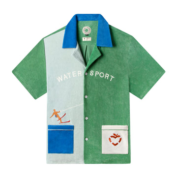 Front view product photo of shirt showing the light blue and green color blocked shirt panels along with the deeper blue collar and left pocket, also featuring the appliquéd waterskier, hotel magique logo life preserver, and embroidered water sport across the chest