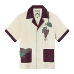 Casual, linen cabana style in pale yellow with eggplant color collar, short sleeves and pockets, Appliqué and edge-embroidered grapes adorn one pocket and the wearer's left chest