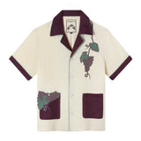 Casual, linen cabana style in pale yellow with eggplant color collar, short sleeves and pockets, Appliqué and edge-embroidered grapes adorn one pocket and the wearer's left chest