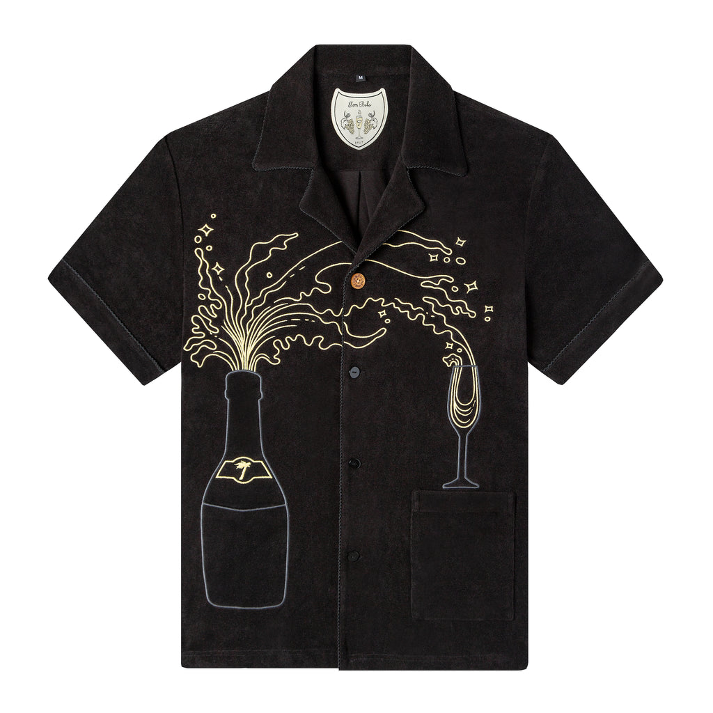 Tombolo Big Brut cabana shirt in Black terry cloth against white background, depicting champagne flowing out of bottle into glass