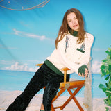 Female model wearing the angler sweater leaning in a chair wearing green pants