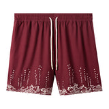 Maroon swim trunks with embroidered fish and mermaids at the leg openings with white bubbles climbing up the legs